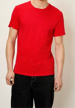 Man wearing plain bright red short-sleeve t-shirt and black pants, standing against a light neutral background.