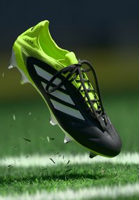 Black and bright yellow soccer cleat with three white stripes, featuring laces, a textured upper, and a streamlined shape, floating above grass.
