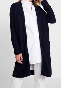 Mujer que lleva un cárdigan de punto azul marino con bolsillos delanteros sobre una camisa blanca de botones y pantalones blancos.