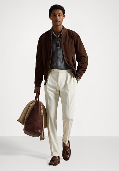 Hnědá semišová bomber bunda, světle šedá košile s knoflíky, krémové kalhoty, hnědé mokasíny, nese hnědou koženou tašku a béžový svetr.