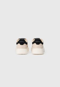 Beige semskinn-sneakers med svarte accenter, med polstret krage, hvit såle og logo-detalj bak. Renslig og moderne design.