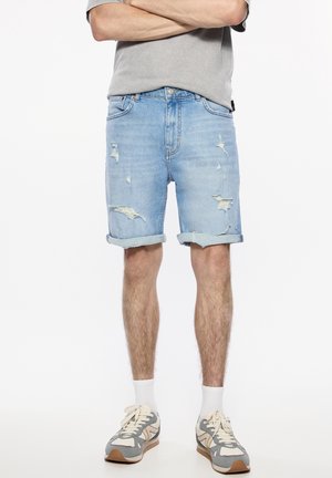 Homme portant un short en jean déchiré bleu clair, un t-shirt gris, des chaussettes blanches et des baskets grises avec des touches crème, debout les bras croisés.
