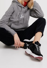 Sudadera gris con un logo, combinada con pantalones de pana negros y zapatillas negras gruesas con acentos en blanco y rojo. Conjunto informal.