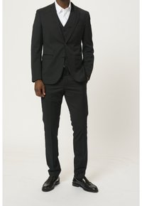 Completo tre pezzi nero con un design aderente, composto da giacca monopetto, pantaloni abbinati e una camicia bianca sotto. Completato con scarpe di pelle nera.
