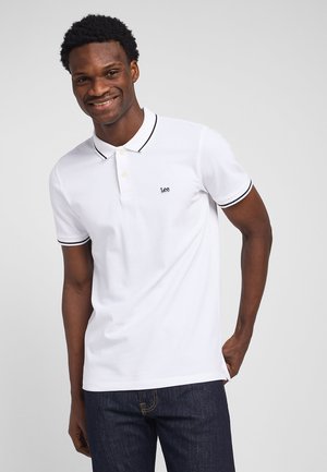 Smilende mand iført hvid polo med sort kant og Lee-logo, stående med den ene hånd i lommen foran en ensfarvet baggrund.