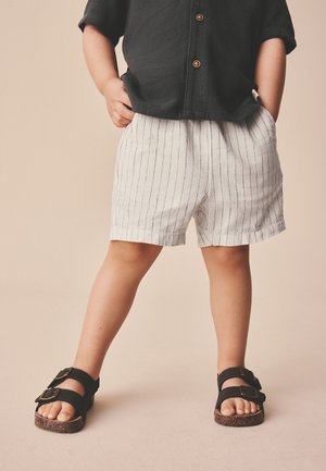 Kind steht mit Händen in den Taschen, trägt ein schwarzes Hemd mit Knöpfen, weiße Shorts mit Nadelstreifen und schwarze Sandalen mit Schnallen auf beigem Hintergrund.
