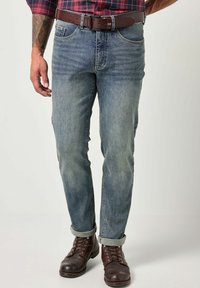 Vaqueros de mezclilla azul con corte recto, textura ligeramente desgastada, puños enrollados y un cinturón de cuero marrón. Combinados con botas marrones de cordones.