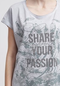 Camisa gris de manga corta con un patrón de hojas, adornada con tachuelas plateadas y el texto "COMPARTE TU PASIÓN" en letras negritas.