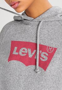 Sudadera gris hecha de material suave, con un gran logo rojo de Levi's en un diseño de arco clásico. Incluye detalles de cordón en la capucha.