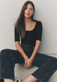 Top nero a coste con maniche corte e dettagli con bottoni, abbinato a jeans scuri a vita alta con un motivo sottile. Scarpe con tacco neutro.