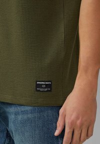 Oliwkowa, fakturowana koszula z krótkim rękawem z czarną metką z napisem "EVOLVING ROOTS". Tkana tkanina w wzór kratkę.