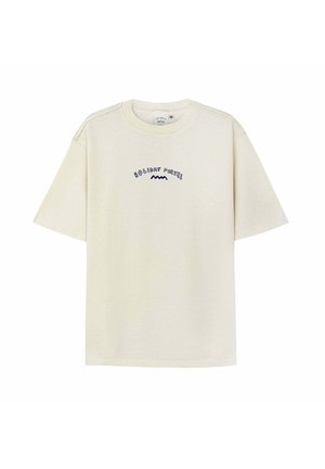 Kobieta z długimi włosami, ubrana w beżową koszulę z graficznym wzorem i napisem „Holiday Cocktail” na plecach, stoi obok czarnego pojazdu.