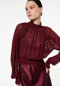 Blusa de encaje burdeos con cuello alto fruncido y puños, que presenta un corte holgado, diseño plisado y textura transparente sobre una falda de satén.