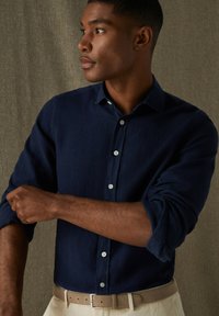 Námornícka košeľa s dlhými rukávmi z ľahkej látky, s rukávmi vyhrnutými pri manžetách. Skombinovaná s béžovým opaskom, na textúrovanom pozadí.