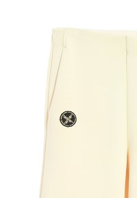 Pantalones beige a medida con pliegues delanteros, que cuentan con un parche redondo negro en el lado izquierdo. Textura de tela suave sin patrones prominentes.