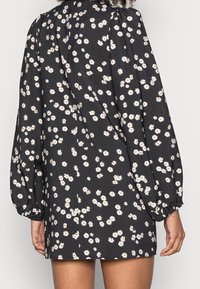 Schwarzes Kleid mit weißem Blumenmuster, das puffärmelige lange Ärmel, einen runden Ausschnitt und einen geraden Schnitt aufweist. Der Stoff wirkt glatt und leicht.