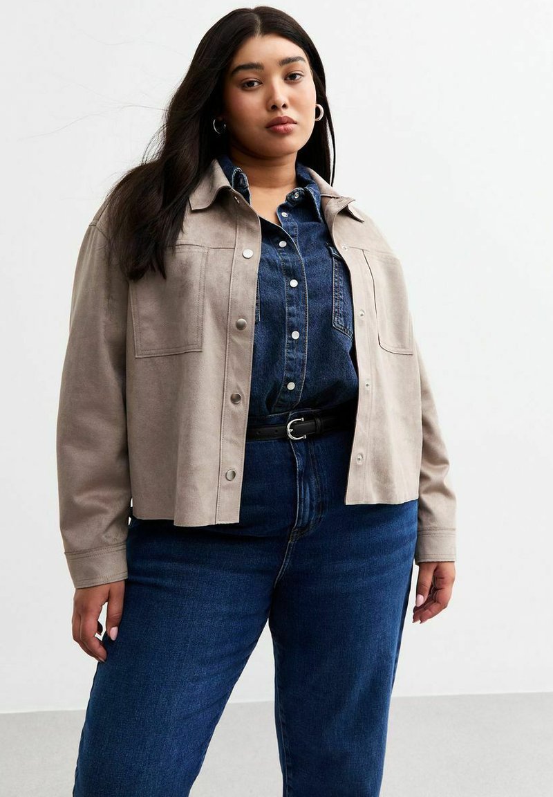 Chaqueta de ante gris con botones a presión, dos bolsillos frontales, llevada sobre una camisa de mezclilla oscura, combinada con jeans azules de talle alto y un cinturón negro.