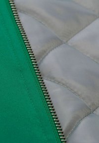 Grüner Stoff mit glatter Textur neben einem hellgrauen gesteppte Material. Enthält einen silbernen Metallreißverschluss entlang der Naht.
