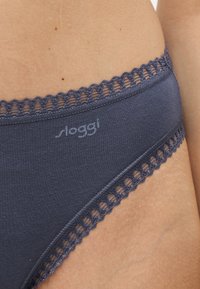 Gros plan d'une personne portant des sous-vêtements gris foncé avec un galon en dentelle et le nom de marque "sloggi" imprimé sur le tissu.