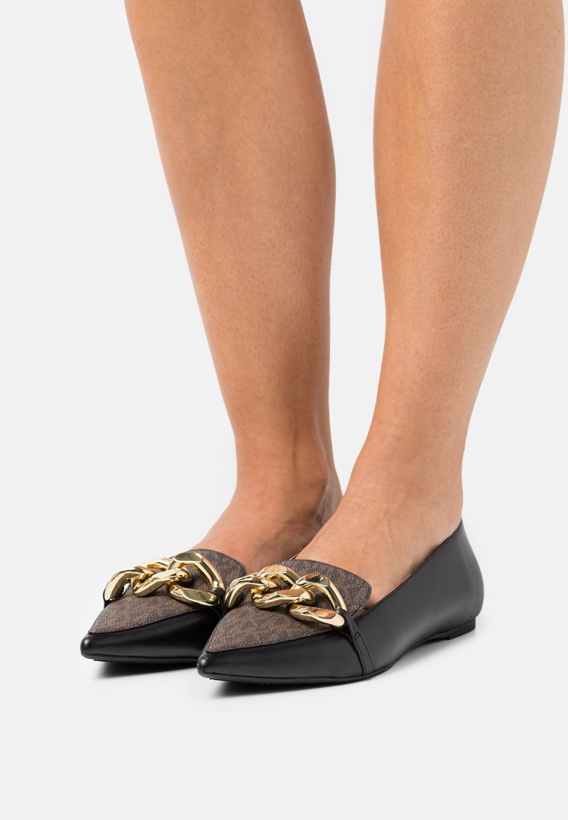 Personne portant des ballerines pointues noires avec des inserts en tissu marron et une grande décoration en chaîne dorée brillante sur fond blanc.
