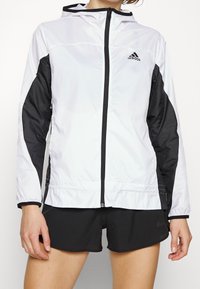 Lehká bílá a černá bunda s kapucí, vybavená předním zipem, elastickými manžetami a hladkým, elegantním povrchem. Viditelné logo Adidas.