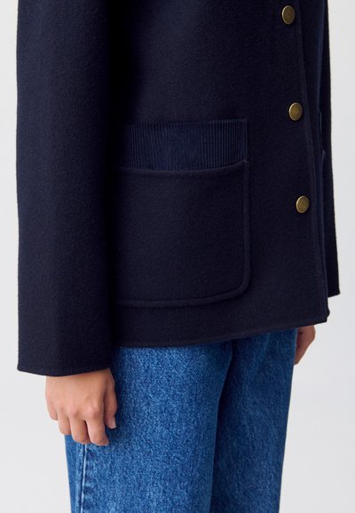 Giacca in misto lana blu navy con tasca in corduroy, caratterizzata da bottoni dorati e una vestibilità relax, abbinata a jeans azzurri chiari.