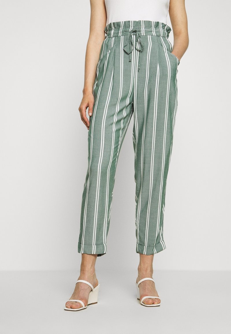 Pantalon rayé vert à coupe décontractée, ceinture élastique, poches latérales et rayures blanches verticales, assorti à des sandales blanches.