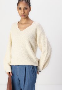 Cream, textured knit sweater with a V-neck and oversized sleeves. Worn with loose blue trousers and accessorized with a brown bag.