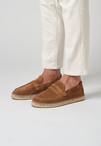 Mocasines de ante marrón con un diseño cosido, que presentan un estilo sin cordones y suela de yute trenzada. Llevados con pantalones cortos blancos.