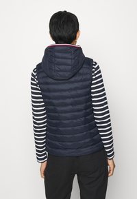 Henkilö, jolla on päällään tummansininen vuorattu huppuliivi musta-valkoisen raidallisen pitkähihaisen paidan päällä, seisoo selin valkoista taustaa vasten.