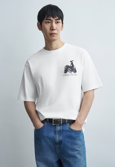 Λευκό βαμβακερό t-shirt με γραφικό σχέδιο στο στήθος, συνδυασμένο με μπλε τζιν τζιν και μαύρη ζώνη, μοντέλο με τα χέρια στις τσέπες.