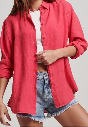 Vrouw draagt een felrode blouse over een wit kort topje, gecombineerd met lichtblauwe, rafelige denim shorts.