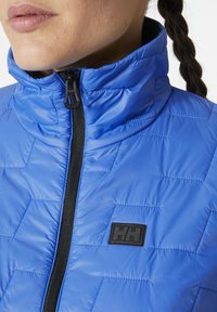 Lehká modrá prošívaná bunda s vysokým límcem, s černým zipem a malou nášivkou na hrudi.