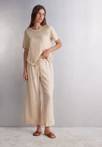 Beige Leinen-T-Shirt mit lockerer Passform, kombiniert mit passenden Jogginghosen mit Kordelzug, getragen mit braunen Slide-Sandalen vor einem hellen Hintergrund.