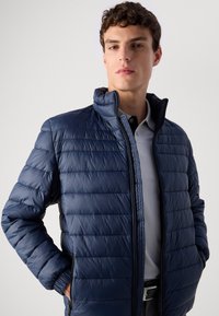 Young man with short curly hair wearing a navy blue quilted jacket over a light gray polo shirt, standing with hands in jacket pockets.