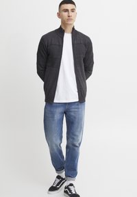 Veste zippée gris foncé avec des panneaux texturés, portée sur un t-shirt blanc, assortie à un jean bleu clair et des baskets noires.
