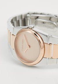 Calvin Klein karóra kerek, rózsaarany számlappal, ezüst szíjjal, kvarc mechanizmussal és minimalista dizájnnal. Ezüst és rózsaarany részletek.