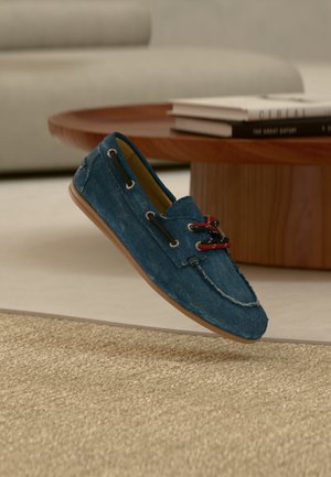 Mocasín casual azul vaquero con suela marrón y cordones rojo-negros flotando sobre una alfombra beige en un salón moderno.