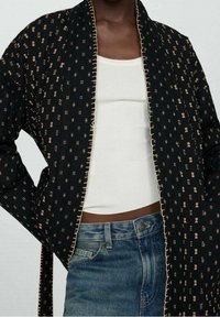 Czarna kurtka w stylu kimona z misternymi złotymi i białymi wzorami, z teksturowanym wykończeniem oraz kontrastowym jasnym szwem wzdłuż krawędzi.