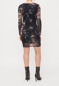 Femme portant une mini-robe noire transparente à fleurs et manches longues, avec des bottines noires à talons hauts, debout de dos.