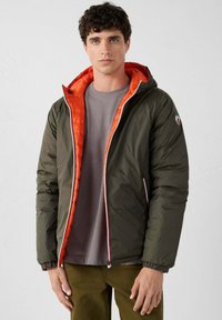 Giacca a vento con un'esterno verde oliva e foderato in arancione. Presenta un cappuccio, chiusura con zip e polsini elasticizzati. Adatta per il clima freddo.