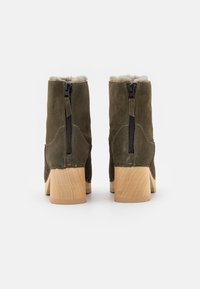 Botines de ante verde oliva con un suave forro de piel, cremalleras traseras y tacones de madera gruesos. Tanto texturizados como distintivos en diseño.
