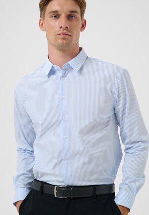 Hombre con camisa de botones a rayas blancas y azules claras metida en pantalones negros con cinturón negro, de pie frente a un fondo liso.