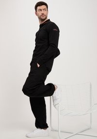 Czarna koszula z długim rękawem i kołnierzykiem, z subtelnym pomarańczowym akcentem. Połączona z czarnymi spodniami i białymi butami sportowymi, oparta na drucianym krześle.