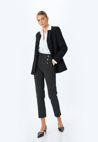 Schwarzer Blazer über einem weißen Hemd, kombiniert mit hochgeschnittenen schwarz-weißen gestreiften Hosen mit Knöpfen. Abgerundet mit schwarzen Absätzen.