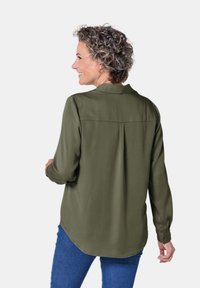 Olivegrüne Bluse mit langen Ärmeln, Kragen und Rückenfalte, kombiniert mit blauen Jeans. Sanfte Textur und lässige Passform.