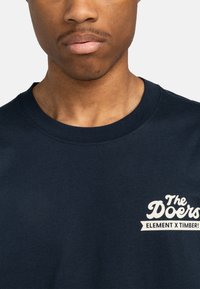 Marineblaues Baumwoll-T-Shirt mit Rundhalsausschnitt; präsentiert ein weißes grafisches Textdesign auf der linken Brust mit der Aufschrift "The Doers ELEMENT X TIMBER!"