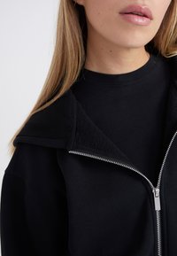 Chaqueta negra con cremallera y un ancho cuello, con una suave textura interior, llevada sobre una camiseta negra a juego.