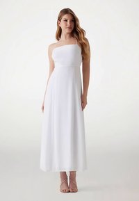 Robe maxi blanche à fines bretelles spaghetti, corsage ajusté et jupe fluide. Associée à des sandales à talons roses clairs. Texture lisse.