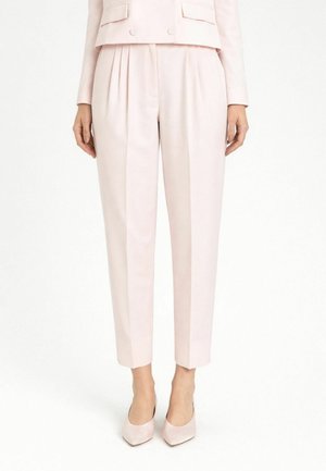 Donna con pantaloni rosa chiaro su misura, corti con pieghe e tacchi a punta abbinati, in piedi su sfondo bianco.